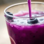Fresh blueberry juice being poured into a glass with a vibrant purple hue, garnished with fresh blueberries and a pink straw.